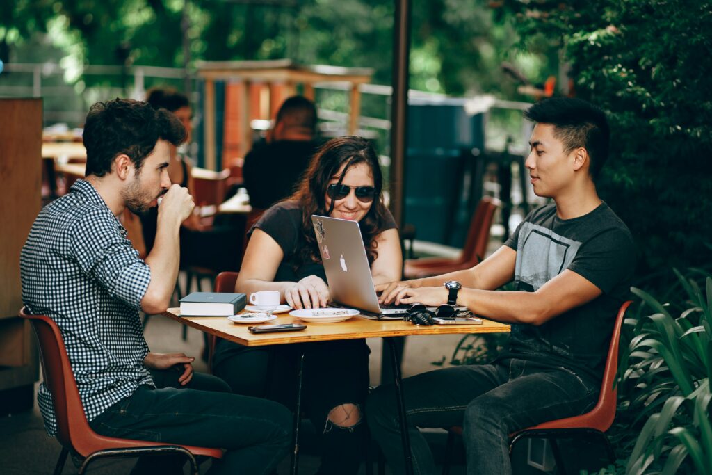 Outsourcing Press Release Distribution is a Game-Changer for PR Professionals 1 a group of people sat around a table at a coffee shop, working together on a laptop discussing Outsourcing press release distribution.